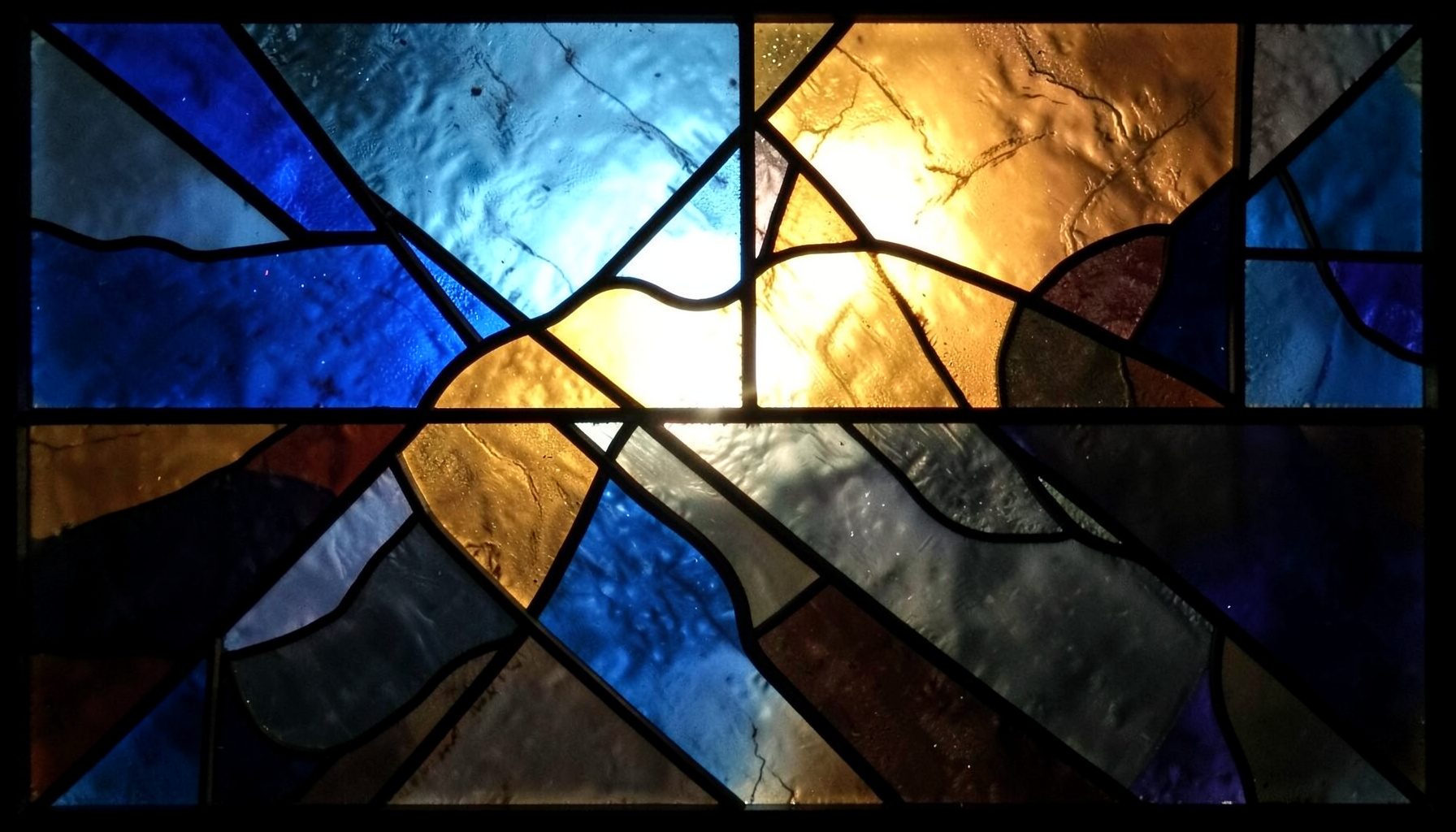 A stained-glass composition with bold leaded segments in deep blues, warm amber, and charcoal tones, forming an abstract geometric pattern that suggests creative craftsmanship and structured thinking